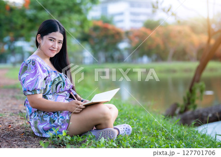 Happy Relaxing Portrait of freelancer asian woman Wear purple dress while working holding diary book writing note while sitting on green grass lawn beside a reservoir at the city park outdoors. 127701706