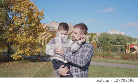 In a bright autumn park, a father gently holds his son, surrounded by colorful foliage and a playground, enjoying quality time on a sunny day. In a bright autumn park, a father gently holds his son, surrounded by colorful foliage and a playground, enjoying quality time on a sunny day. 127702590