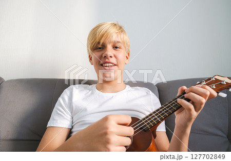 Caucasian Boy Playing Ukulele while Sitting on Sofa, Looking into Camera. Young Musician Learning to Play Instrument at Home. Caucasian Boy Playing Ukulele while Sitting on Sofa, Looking into Camera. Young Musician Learning to Play Instrument at Home. 127702849
