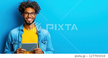 Young man with curly hair stands against a blue background holding a tablet with a cheerful expression during daytime 127703410