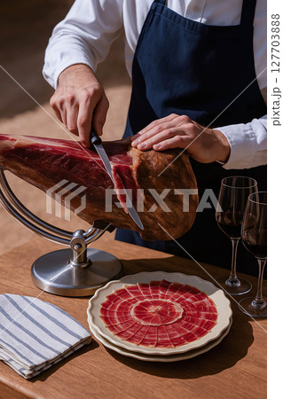 A gourmet scene showing a man in a white shirt and navy apron skillfully slicing a leg of  ham 127703888