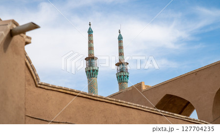 Intricately tiled minarets of Jameh Mosque of Yazd, rising above traditional mud-brick buildings under a sunny sky in Yazd Intricately tiled minarets of Jameh Mosque of Yazd, rising above traditional mud-brick buildings under a sunny sky in Yazd 127704235