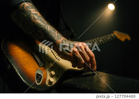 Tattooed rock guitarist adjusting vintage sunburst electric instrument under moody spotlight captures raw energy passion of live performance stage 127704480