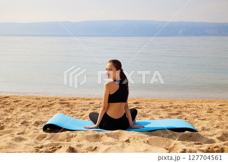 A sporty girl is sitting on a yoga mat on a sandy beach on the shore of a lake. Yoga time. 127704561