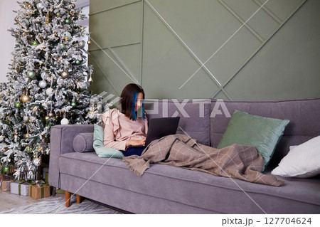 A pretty girl is working on a laptop, covered with a blanket. Christmas tree with toys and gifts. 127704624