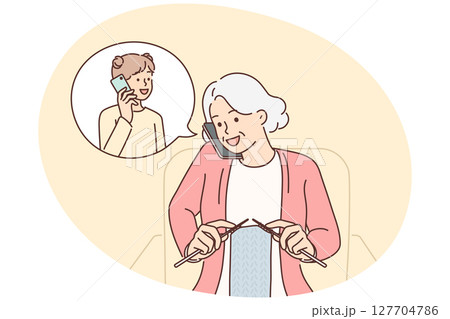 Grandmother makes phone call to granddaughter and knits scarf sitting in cozy chair 127704786