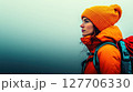 Profile of a woman in an orange jacket and yellow beanie hiking in a foggy mountain landscape during early morning hours 127706330