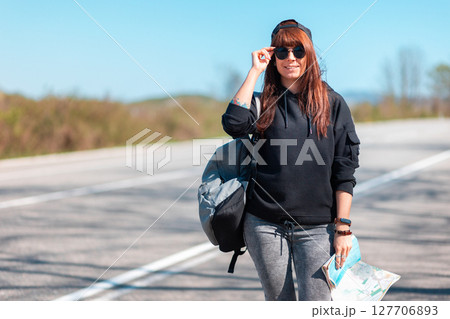 Concept of hitchhiking and local travel. A hipster happy girl in cap and sunglasses with tattoed hand stand along the road with a paper map. Copy space 127706893