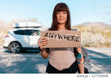 Portrait of pretty smiling caucasian young woman with tattoed hand holding a cardboard sign with text anywhere. Car on the background. The concept of local traveling and hitchhiking Portrait of pretty smiling caucasian young woman with tattoed hand holding a cardboard sign with text anywhere. Car on the background. The concept of local traveling and hitchhiking 127706896