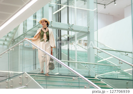 Wide view of a Caucasian young elegant woman in a straw hat and carrying a bag descending transparent glass steps. In the background is a modern glass business building 127706939