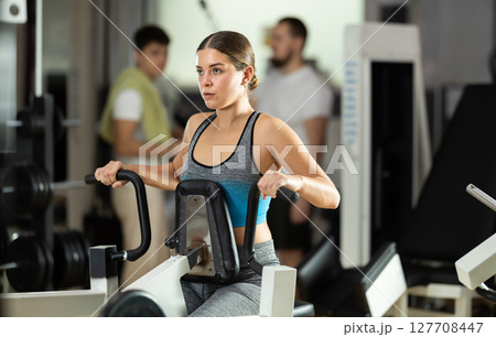 Girl in gym performs exercises for arm muscles using simulators and fitness center equipment. Girl in gym performs exercises for arm muscles using simulators and fitness center equipment. 127708447