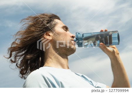 Handsome brunette with long hair drinking water from bottle in sunny day against clear blue sky. Summer. Handsome brunette with long hair drinking water from bottle in sunny day against clear blue sky. Summer. 127709994