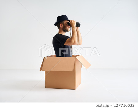 Man in hat looking through binoculars sitting inside cardboard box. 127710032