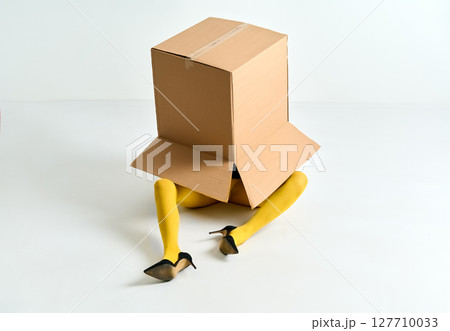 Woman in yellow tights and black high heels hidden under oversized cardboard box on white floor 127710033