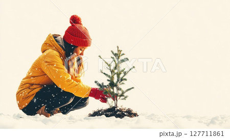 Young girl plants a small tree in the snow during winter while wearing a bright yellow coat and red hat Young girl plants a small tree in the snow during winter while wearing a bright yellow coat and red hat 127711861