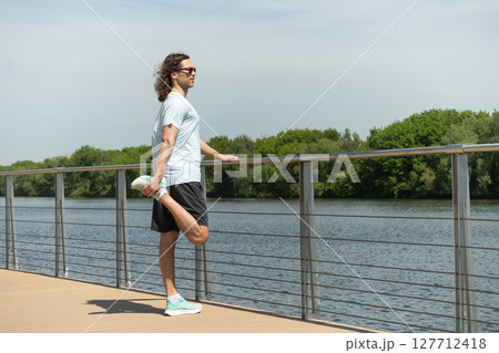 Fit handsome brunette man wearing sports casual clothes doing stretching legs exercise on city outdoor public park. Summer activity. Healthy lifestyle. 127712418