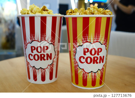 Two large striped red, yellow snd white popcorn paper cups with the "POPCORN" text. Salty popcorn and caramel popcorn in paper cups on table in cinema hall Two large striped red, yellow snd white popcorn paper cups with the "POPCORN" text. Salty popcorn and caramel popcorn in paper cups on table in cinema hall 127713676