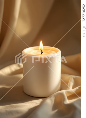A single white candle burning brightly on a textured beige fabric surface, creating a peaceful and warm light. A single white candle burning brightly on a textured beige fabric surface, creating a peaceful and warm light. 127716252