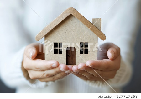Wooden miniature house held gently in hands against a soft background, symbolizing home and warmth in a cozy indoor setting 127717268