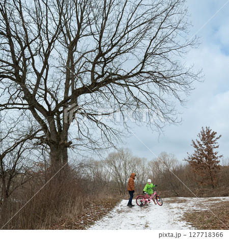 Winter adventure unfolds as a child learns to ride a bicycle near a majestic old tree in a serene, snowy landscape during a quiet afternoon 127718366
