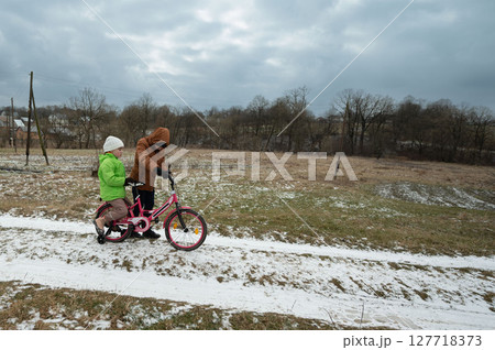 Children enjoying a winter day in a snowy landscape while riding a colorful bicycle along a grassy path under an overcast sky Children enjoying a winter day in a snowy landscape while riding a colorful bicycle along a grassy path under an overcast sky 127718373