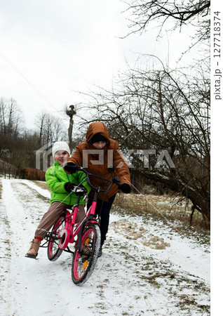 Two joyful children riding a bright pink bicycle together on a snowy path in winter, surrounded by barren trees and a tranquil landscape Two joyful children riding a bright pink bicycle together on a snowy path in winter, surrounded by barren trees and a tranquil landscape 127718374