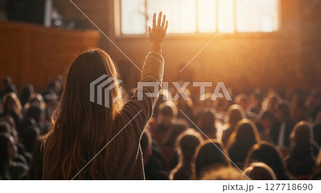 A person with long hair raises their hand in a crowd during a worship or concert gathering, backlit by warm golden sunlight streaming through windows 127718690