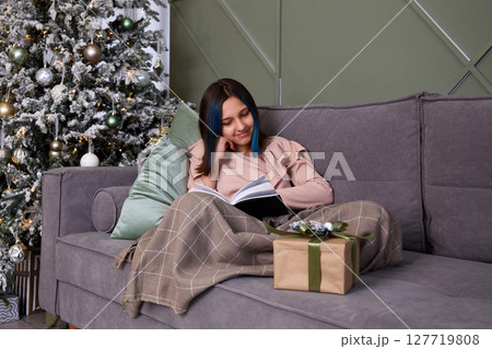 A cute girl is reading a book on the sofa, covered with a blanket. Next to the Christmas tree with toys and gifts. A cute girl is reading a book on the sofa, covered with a blanket. Next to the Christmas tree with toys and gifts. 127719808