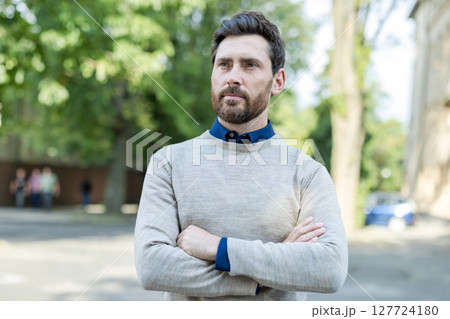Confident adult male professional standing with arms crossed on urban street. Mature businessman exudes leadership and focus, dressed in casual attire. Outdoors setting highlights city environment 127724180