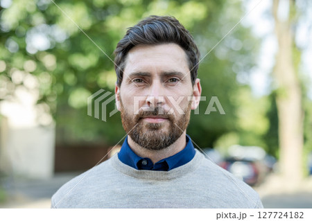 Portrait of confident man with beard outdoors wearing casual sweater and blue shirt. Expression of determination and calmness. Perfect for themes of fashion, lifestyle, or professionalism. 127724182