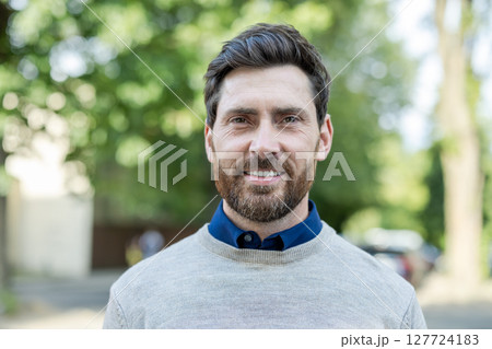 Mature man with beard stands confidently outdoors wearing a sweater and shirt. Smiling businessman presents a friendly, approachable demeanor ideal for business settings. Mature man with beard stands confidently outdoors wearing a sweater and shirt. Smiling businessman presents a friendly, approachable demeanor ideal for business settings. 127724183