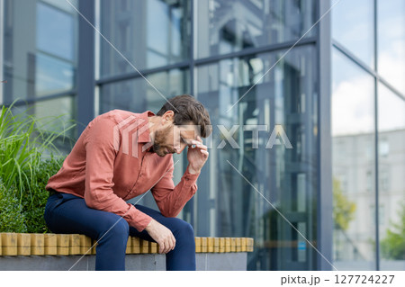 Mature businessman in city street sitting outside office building, showing stress and contemplation. Professional adult expressing concern about work, dressed casual, highlighting urban lifestyle. 127724227