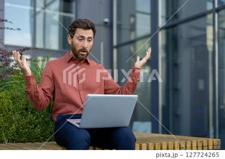 Mature man sitting outdoors in urban setting, using laptop with surprised expression. Wearing casual attire, he reacts to unexpected online news or communication, illustrating shock and emotion. Mature man sitting outdoors in urban setting, using laptop with surprised expression. Wearing casual attire, he reacts to unexpected online news or communication, illustrating shock and emotion. 127724265