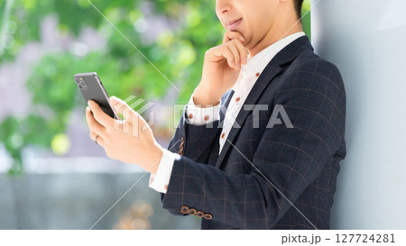 スマートフォンの画面を見て考える男性ビジネスマン スマートフォンの画面を見て考える男性ビジネスマン 127724281