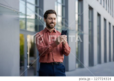 Mature male professional standing outside in city setting, holding phone. Man wearing casual attire, engaging with smartphone. Depicts business, communication, technology, and lifestyle concepts. 127724314