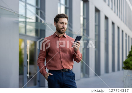 A man in a red shirt is looking at his cell phone. He is standing in front of a building A man in a red shirt is looking at his cell phone. He is standing in front of a building 127724317