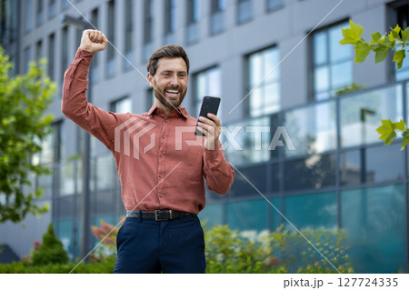 Joyful businessman looking at smartphone celebrates success on city street. Mature professional expressing excitement outside modern office building, representing success, achievement Joyful businessman looking at smartphone celebrates success on city street. Mature professional expressing excitement outside modern office building, representing success, achievement 127724335