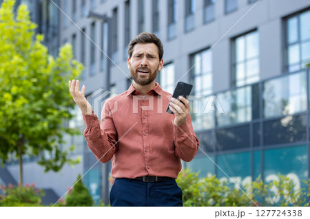 A man in a red shirt is holding a cell phone. He is looking at the phone and he is upset A man in a red shirt is holding a cell phone. He is looking at the phone and he is upset 127724338