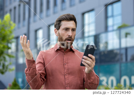 Mature businessman standing on city street holding phone with shocked expression. Wearing casual red shirt, adult man reacts to surprising message. Outdoors setting highlights urban work environment. Mature businessman standing on city street holding phone with shocked expression. Wearing casual red shirt, adult man reacts to surprising message. Outdoors setting highlights urban work environment. 127724340