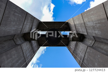 A large dark bell hangs prominently silhouetted within a monumental brutalist concrete structure, viewed from below against a vibrant blue sky, conveying remembrance and history A large dark bell hangs prominently silhouetted within a monumental brutalist concrete structure, viewed from below against a vibrant blue sky, conveying remembrance and history 127724413