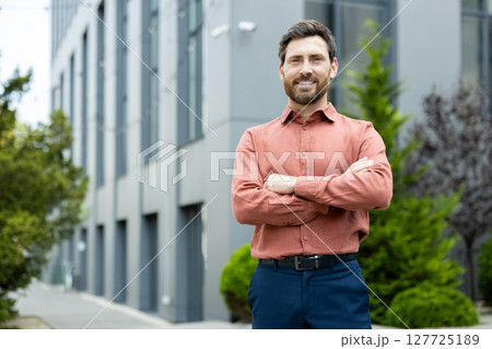 Portrait of successful businessman outside office building, man in shirt smiling and looking at camera with crossed arms, satisfied office worker in casual clothes. 127725189