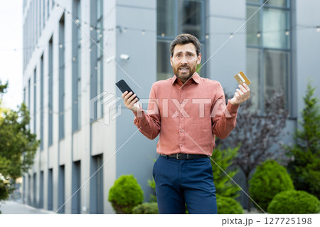 Portrait of a deceived and disappointed man, holding a bank credit card and phone in his hands, account blocked, and money transfer error Portrait of a deceived and disappointed man, holding a bank credit card and phone in his hands, account blocked, and money transfer error 127725198
