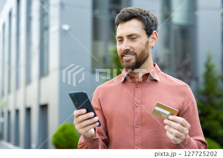 A man smiling while holding a credit card and phone, likely making an online purchase outdoors. A man smiling while holding a credit card and phone, likely making an online purchase outdoors. 127725202