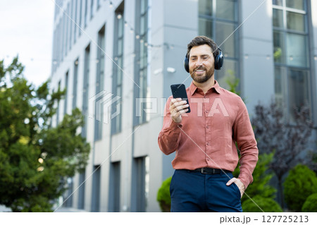 A man wearing headphones listens to music on his smartphone outdoors near an office building, enjoying a relaxing moment. 127725213