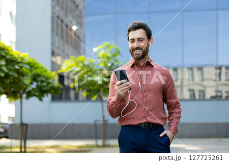 A well-dressed man listens to music or communicates on his phone while outdoors. The sunlight catches his smiling face as he enjoys a moment. 127725261