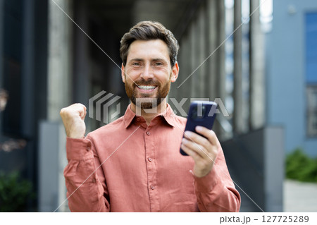 A man with a beard wearing a button-down shirt is holding a smartphone and celebrating success, outdoors with an urban background. 127725289