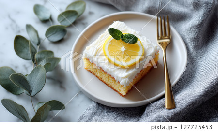 Piece of lemon cake with white frosting, garnished with a lemon slice and mint, served on a white plate with a gold fork. Elegant and fresh dessert. Citrus flavor. 127725453