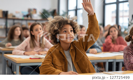Curly-haired schoolboy raising his hand in a classroom during a lesson, surrounded by attentive classmates. Active participation in education. Bright learning moment. 127725499