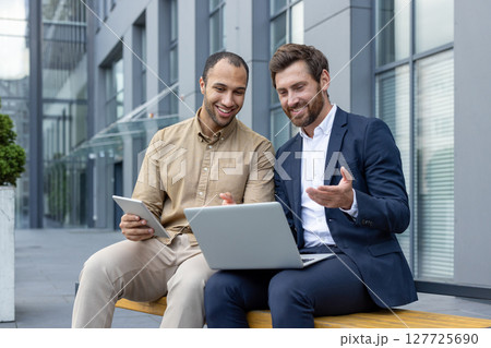 Two business partners sit outdoors, collaborating on a laptop and tablet, discussing work-related data. 127725690