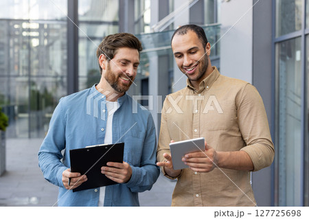 Two diverse businessmen review a tablet, possibly discussing a project or presenting ideas, smiling in a modern outdoor setting. Two diverse businessmen review a tablet, possibly discussing a project or presenting ideas, smiling in a modern outdoor setting. 127725698